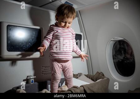 Verletzung der Sicherheitsregeln im Flugzeug. Gefahr. Niedliches kleines Kleinkind steht während des Fluges in Babybettchen. Stockfoto