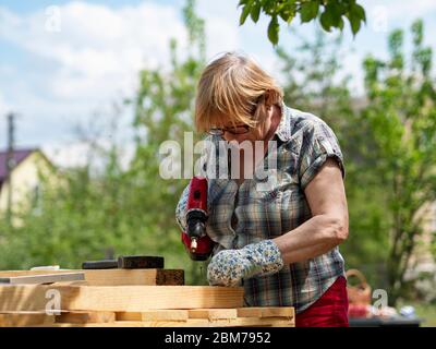 Ältere kaukasische Frau montiert einen Holzrahmen eines Gebäudes mit Metallbefestigungen mit einem Schraubendreher Stockfoto