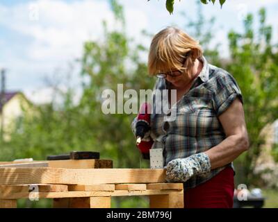 Ältere kaukasische Frau montiert einen Holzrahmen eines Gebäudes mit Metallbefestigungen mit einem Schraubendreher Stockfoto