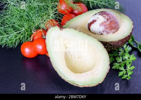 Ketogene Diät Avocado. Hälften Avocado, Tomaten Kirsche und frischen Dill mit Petersilie auf dem schwarzen Küchenbrett. Gesunde ketogene Diät Lebensmittel. Stockfoto