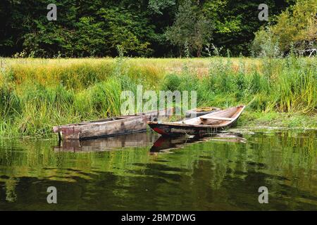 Zwei alte Boote am Ufer sind an ein altes Autorad gebunden, auf einem grünen Grasgrund ein sonniger Sommertag Stockfoto