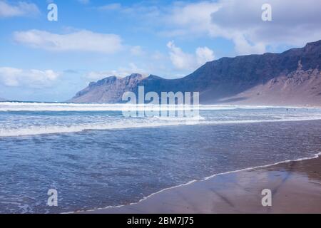 Wellen umrunden den Strand mit einer spektakulären Klippenkulisse in der beliebten Wassersportstation Caleta de Famara auf Lanzarote, Kanarische Inseln Stockfoto