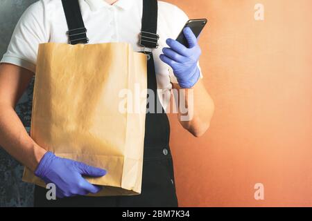 Kurier in Uniform und medizinische Handschuhe mit Papiertüte mit Lebensmitteln. Volunteer Mann an der Tür liefern Online-Bestellung und überprüfen Telefon. Kontaktlos Stockfoto