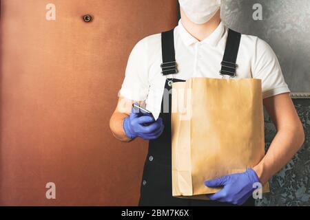 Männliche Kurier in chirurgischen Maske und medizinische Handschuhe mit Papiertüte mit Essen zum Mitnehmen. Volunteer Mann an der Tür liefern Online-Bestellung und überprüfen Telefon. Stockfoto