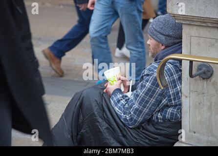 Ein Obdachloser sitzt in einem Schlafsack und hält einen Becher für Spenden am Eingang zur U-Bahn-Station Oxford Circus im Londoner West End Stockfoto