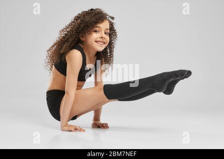 Seitenansicht der kleinen weiblichen Profimartin, die auf Armen mit Beinen auf dem Boden steht, isoliert auf grauem Hintergrund. Lächelndes Mädchen in Sportswear und Kniestrümpfe mit lockigen Haaren zeigt Flexibilität. Stockfoto