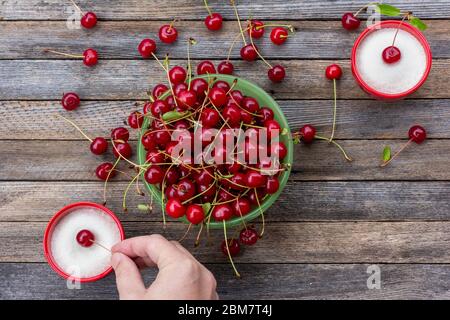 Eine grüne Schale mit reifen Kirschbeeren und einer männlichen Hand taucht eine Kirschenbeere in eine Tasse mit Zucker auf einen Holztisch, Nahaufnahme Draufsicht Stockfoto