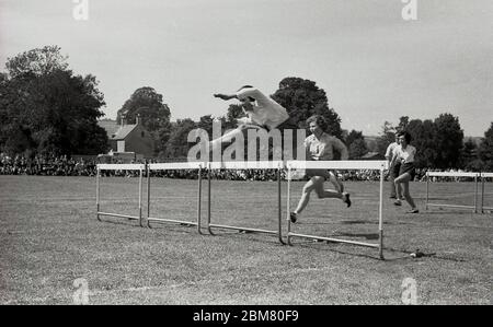 Sekundarschulsport, England, c60s, draußen auf einer Grasbahn, Teenager-Mädchen in einem Hürdenlauf konkurrieren, mit einem Mädchen an der Vorderseite springen über eine Barriere oder Hürde zeigt gute Technik. Stockfoto