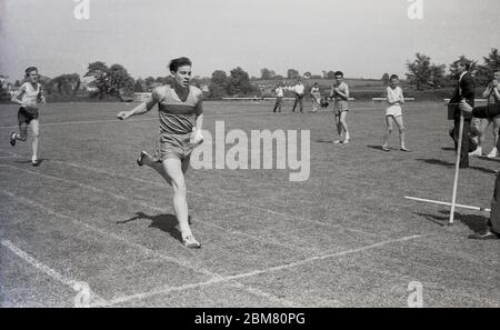 Sekundarschulsport, England, c60s, draußen auf einer Grasbahn, Senioren im Wettbewerb in der 100 Yard Dash oder Sprint Rennen, Bild zeigt einen athletischen jungen Mann im Begriff, das Endband zu schlagen und das Rennen zu gewinnen. Stockfoto