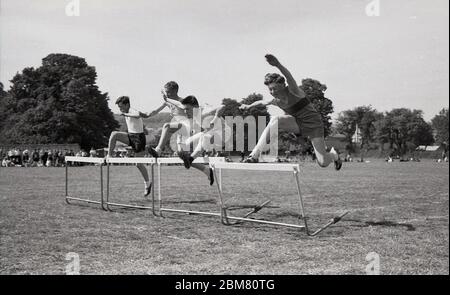 Sporttag der Sekundarstufe, England, c1960s, Leichtathletik, vier Schüler, die bei einem Hürdenrennen draußen auf einer Grasbahn gegeneinander antreten und über die Barrieren springen. Stockfoto