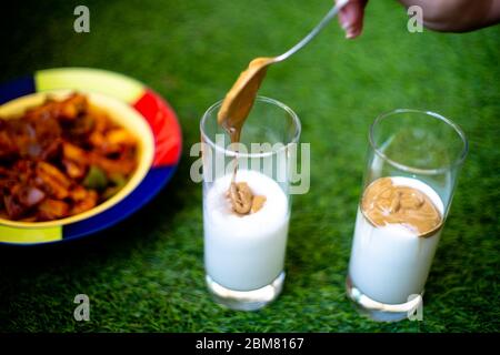 Junge indische Frau mit einem Löffel, um Schlagkaffee auf dicke Sahne Milch zu Dalgona Kaffee in transparentem Glas auf grünem Gras zu machen Stockfoto