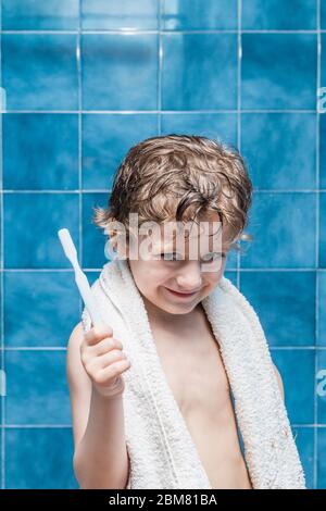 Ein Kind mit einem Handtuch auf den Schultern, das eine Zahnbürste zeigt, mit blauen Fliesen im Hintergrund. Stockfoto