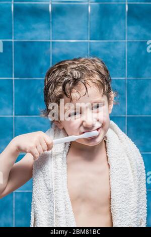 Ein Kind mit einem Handtuch auf den Schultern, das seine Zähne putzt, mit blauen Fliesen im Hintergrund. Stockfoto