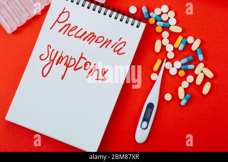 Notizblock, eine Vielzahl von Medikamenten, Thermometer, Spritze und Ampullen mit Medizin auf rotem Hintergrund. Geschrieben in blau auf einem Notizblock Lungenentzündung Symtoms . Stockfoto