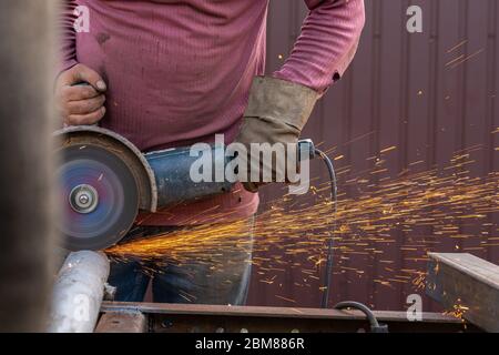 Nahaufnahme an den Seiten fliegen helle Funken aus dem Schweißgerät. Ein junger Schweißmann in Arbeitshandschuhen schleift ein Metallprodukt mit Winkelschleifer ein Stockfoto