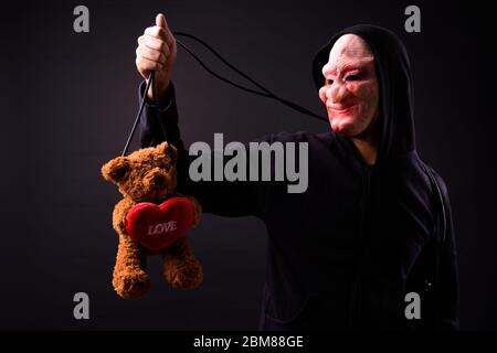 Junger Mann in Kapuzenpullover und Horror Maske hängender Teddybär mit Schnur Stockfoto