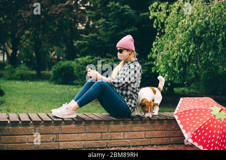 Junges Mädchen mit Schutzmaske mit Smartphone und sitzt auf dem pench im grünen Park mit Hund Stockfoto