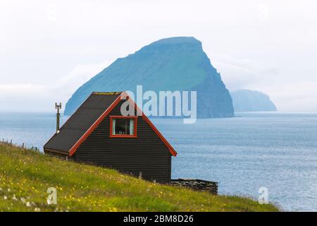 Schwarzes Haus auf dem berühmten färöischen Hexen Finger Trail und der Koltur Insel im Hintergrund. Dorf Sandavagur, Insel Vagar, Inseln der Färöer, Dänemark. Landschaftsfotografie Stockfoto