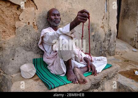 Muslimischer Mann, der auf einer Matte auf dem Boden sitzt und mit einem Rosenkranz in der Hand auf den Straßen von Kano betet. Stockfoto