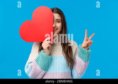 Taille-up-Porträt hübsche weibliche junge romantische Frau, große niedliche rote Herzkarte am Valentinstag, halten Sie sie über Gesicht, zeigen Sie ein Friedenszeichen und lächeln Stockfoto