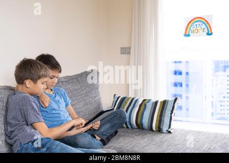 Junge mit Tablet-Computer im Zimmer sitzen. Auf dem Fenster Zeichnung Bild mit Inschrift zu Hause bleiben Stockfoto