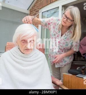 Cocoid 19 Einschränkungen bedeutet, dass die alte Dame nicht wie üblich in den Friseur genommen werden kann, so dass ihre aufsichtschenende Pflegekraft ihr Haar für sie als es ge trimmt und kämmt Stockfoto