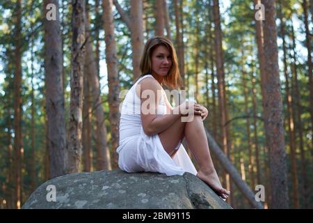 Ein Mädchen in einem weißen Kleid sitzt auf einem Felsen im Wald bei Sonnenuntergang Stockfoto