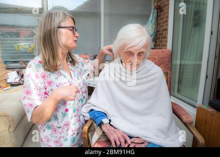 Cocoid 19 Einschränkungen bedeutet, dass die alte Dame nicht wie üblich in den Friseur genommen werden kann, so dass ihre aufsichtschenende Pflegekraft ihr Haar für sie als es ge trimmt und kämmt Stockfoto