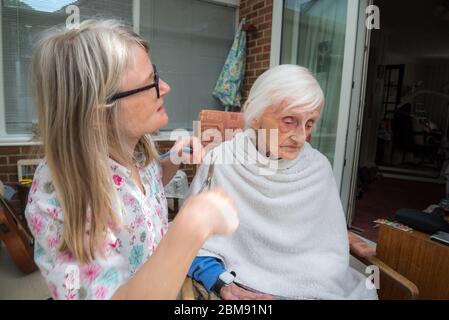 Cocoid 19 Einschränkungen bedeutet, dass die alte Dame nicht wie üblich in den Friseur genommen werden kann, so dass ihre aufsichtschenende Pflegekraft ihr Haar für sie als es ge trimmt und kämmt Stockfoto