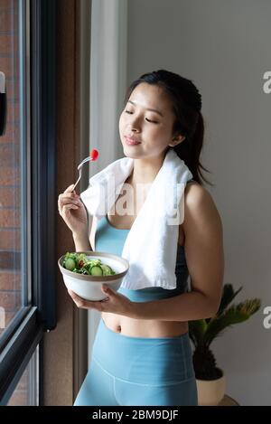 Eine junge asiatische Frau, die gesundes grünes Gemüse am Fenster isst