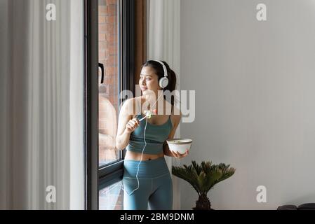 Eine junge asiatische Frau, die gesundes grünes Gemüse am Fenster isst