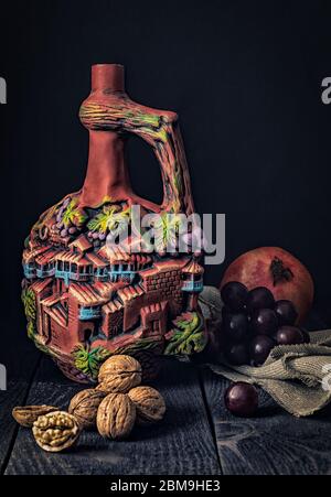 Auf einem Holztisch befindet sich eine Steingutflasche mit einem Krug roten georgischen Kinzmarauli-Wein, umgeben von Trauben, Granatäpfel und Walnüssen. Stillleben. Stockfoto