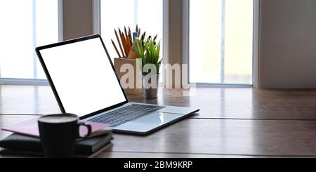 Laptop mit weißem, leeren Bildschirm, der auf einem hölzernen Schreibtisch steht und umgeben von Notizbuch, Kaffeetasse, Topfpflanze, Bleistifthalter und Markierungsstiften. Ord Stockfoto