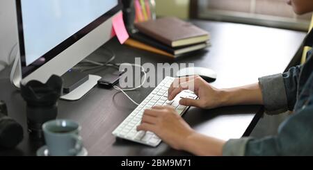 Seitenbild des Mannes, der als Grafikdesign arbeitet, während er am schwarzen Schreibtisch sitzt und auf seiner Computertastatur über dem komfortablen Wohnzimmer A tippt Stockfoto