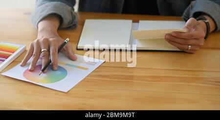 Zugeschnittenes Bild von den Händen des Designers, die einen Bleistift und Papier halten, während sie vor dem Buntstift sitzen, Farbführer und Farbkarte am hölzernen Schreibtisch Ove Stockfoto