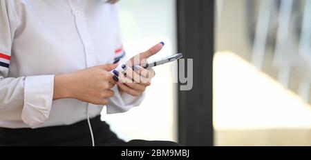 Zugeschnittenes Bild von schönen Frau in weißem Hemd halten / mit Smartphone beim Laden und sitzen über komfortable Wohnzimmer als Hintergrund. Woma Stockfoto