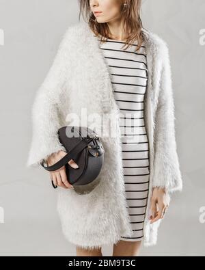Stilvolle modische weiße Frau mit trendigen Leder rund schwarzen Handtasche Tasche. Modell mit weißer flauschiger Strickjacke und gestreiftem Kleid. Kleidung und Zugang Stockfoto
