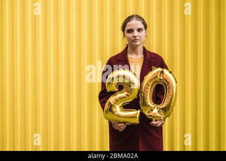 Junge schöne traurige Mädchen in Burgunder Mantel und Senf Pullover hält in ihren Händen die Helium Ballons der Nummer 20 vor dem Hintergrund der hellen yel Stockfoto