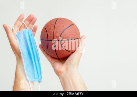 Basketball-Ball mit medizinischen Schutzmaske in den Händen des Mannes auf hellem Hintergrund während Coronavirus mit Kopieraum. Basketball auf Pause wegen Coronavirus. Stockfoto