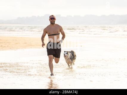Sportmann mit Hunden und Training mit Herzfrequenzsensor am hundefreundlichen Strand. Outdoor-Training und Joggen mit Ihrem besten Freund. Gesund OW Stockfoto