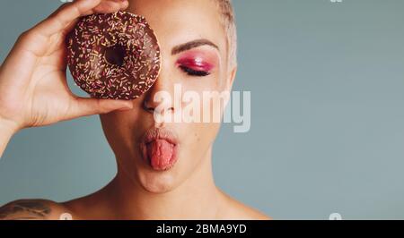 Nahaufnahme einer Frau mit kurzen Haaren, die einen Donut hält und eine herausstehende Zunge hat. Weibliches Model ein Donut vor ihrem Gesicht auf grauem Hintergrund. Stockfoto