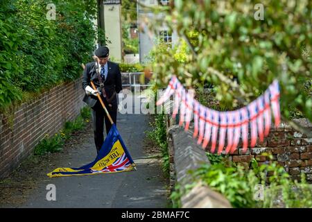 Der Fahnenträger der Royal British Legion Richard Hignet senkt den Standard während der zwei Minuten Stille vor seinem Haus in Royal Wootton Bassett, Wiltshire, während er während eines Veranstaltungstages zum 75. Jahrestag des VE Day sozial distanziert bleibt. Stockfoto