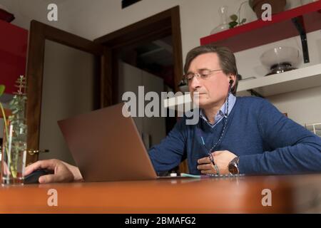 Fünfzig Jahre alter Mann mit Kopfhörern arbeitet von zu Hause aus in der Küche und mit Laptop, neue normal Stockfoto
