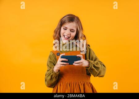 Bild von einem netten emotionalen glücklich jungen Mädchen Kind isoliert über gelbe Wand Hintergrund spielen Spiele per Handy. Stockfoto