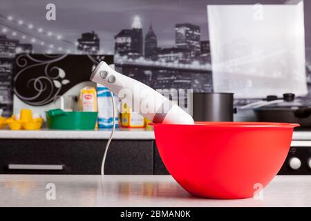 Tauchmixer in einer roten Schüssel auf einem Tisch Nahaufnahme isoliert auf dem Hintergrund von Küchenutensilien und Zutaten für die Herstellung von Teig zum Backen. Stockfoto