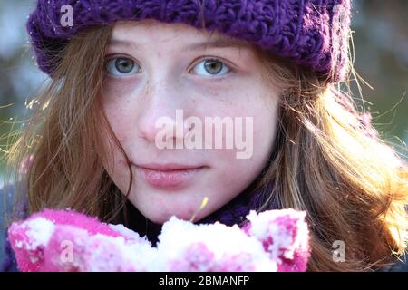 Junge, blauäugige Mädchen mit violettem Strickmütze und rosa Handschuhen, die im Winter Schnee halten und Sonne auf ihrem Haar in Lancaster County, Pennsylvania, scheint Stockfoto