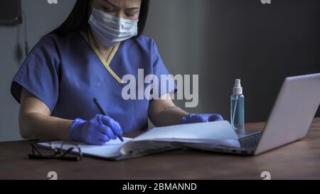 Müde Ärztin füllt medizinische Formulare aus, Frau schreibt mit einem Stift auf Papier sitzt am Schreibtisch, während die Arbeit am Computer in ihrem Büro Stockfoto