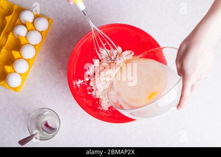 Weibliche Hände halten einen Schneebesen in einer Hand und mit anderen Gießen Schlageier in transparente Glasschüssel in die Quark Masse in roten Teller bereiten Teig für die Stockfoto