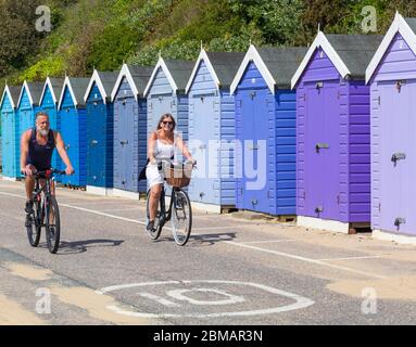 Bournemouth, Dorset, Großbritannien. Mai 2020. UK Wetter: Schöner warmer sonniger Tag mit steigenden Temperaturen am Feiertag Freitag, der VE Tag 75. Jahrestag markiert. Die Strände sind hauptsächlich verlassen, und die Leute nehmen ihre zulässige Bewegung am Meer, die meisten halten sich an die Coronavirus Richtlinien, da Besucher geraten werden, weg zu bleiben und zu Hause zu bleiben. Mann und Frau Radfahren entlang der Promenade vorbei an Strandhütten - Reiten Fahrräder Fahrrad Fahrrad Radfahrer. Quelle: Carolyn Jenkins/Alamy Live News Stockfoto