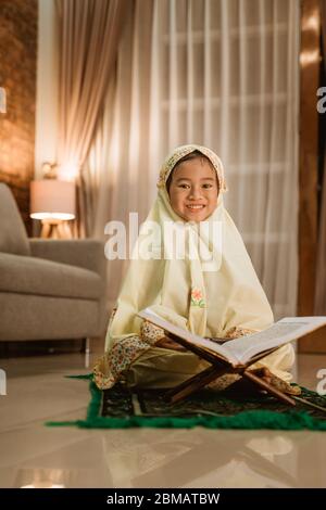 Schönes kleines Mädchen, das den koran liest und allein zu Hause einen muslimischen Hijab trägt Stockfoto